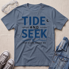 A stone blue short-sleeve graphic T-shirt laid flat on a light beige textured surface, with safety glasses, gray sneakers, and a folded denim jacket around it. The shirt has large navy text reading 'TIDE AND SEEK' with smaller text 'THE MOON FOUND ME' and a crescent moon graphic.