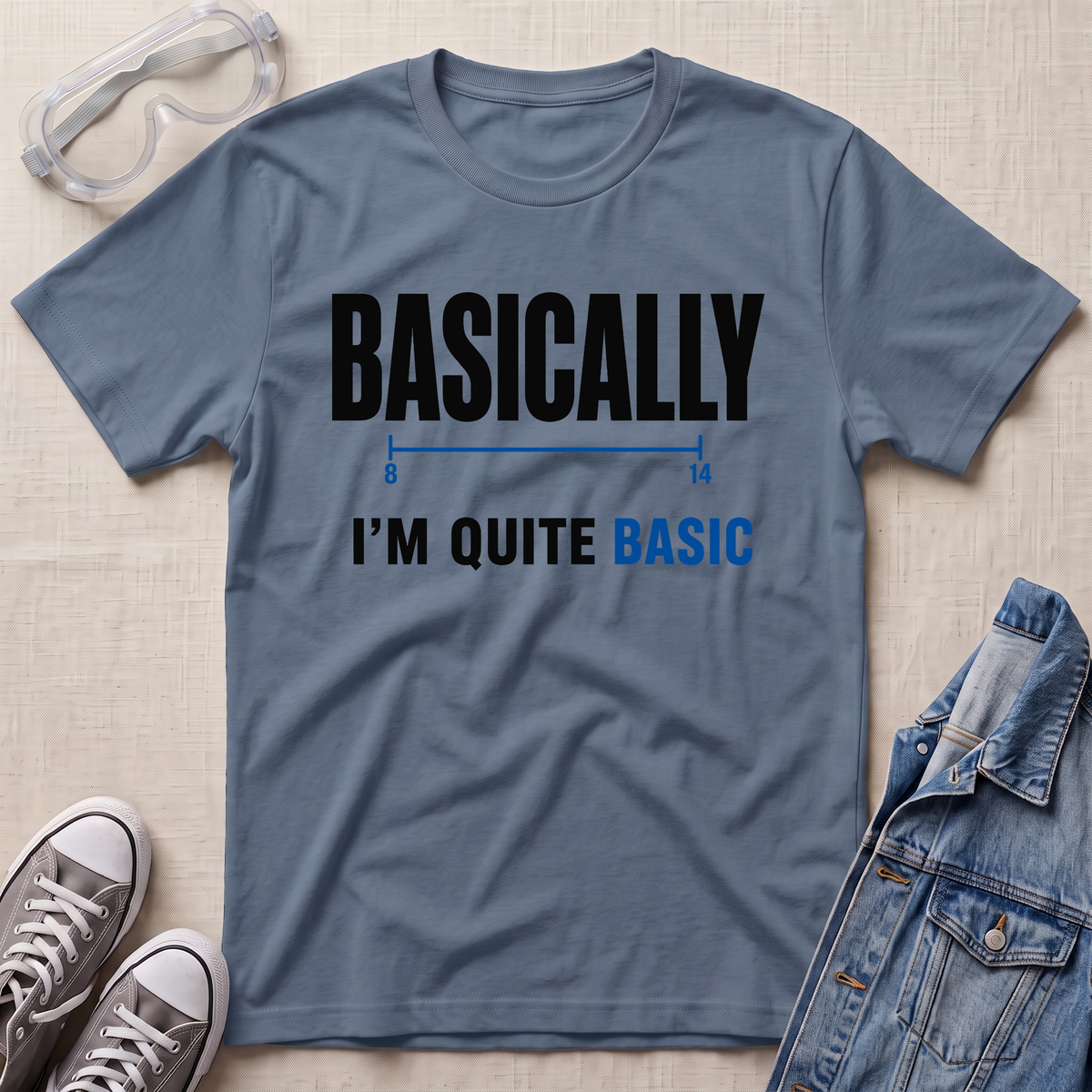A slate blue graphic t-shirt laid flat on a light beige textured background, styled with safety goggles, gray sneakers, and a denim jacket. The t-shirt features the text 'BASICALLY' in black, followed by a blue number line graphic, and 'I'M QUITE BASIC' in black and blue.