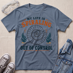A slate blue graphic t-shirt laid flat on a light beige textured background, surrounded by styling props including clear safety glasses, gray sneakers, and a denim jacket. The t-shirt features a graphic of a spiral ammonite shell with the text 'MY LIFE IS SPIRALING OUT OF CONTROL' in orange and dark teal.