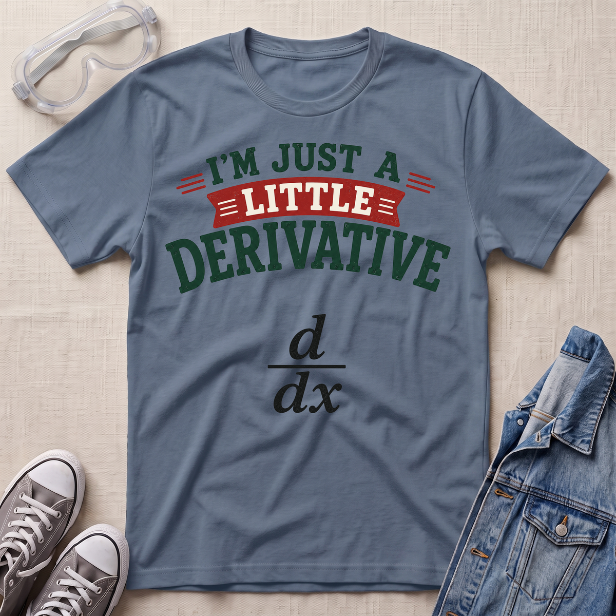 A slate blue graphic t-shirt laid flat on a beige textured background, styled with safety glasses, gray sneakers, and a denim jacket. The shirt features the text 'I'M JUST A LITTLE DERIVATIVE' in green and red, with a black derivative symbol printed below.