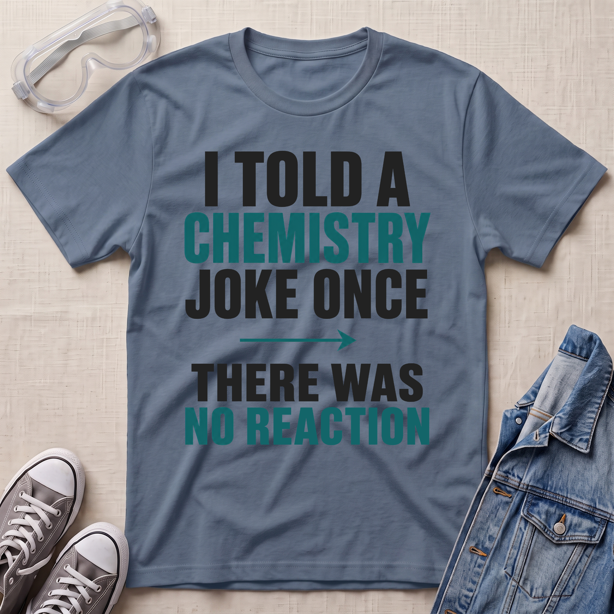 A slate blue graphic t-shirt laid flat on a beige textured background, surrounded by styling props including safety goggles, gray sneakers, and a denim jacket. The t-shirt features the text 'I TOLD A CHEMISTRY JOKE ONCE THERE WAS NO REACTION' in black and teal lettering.