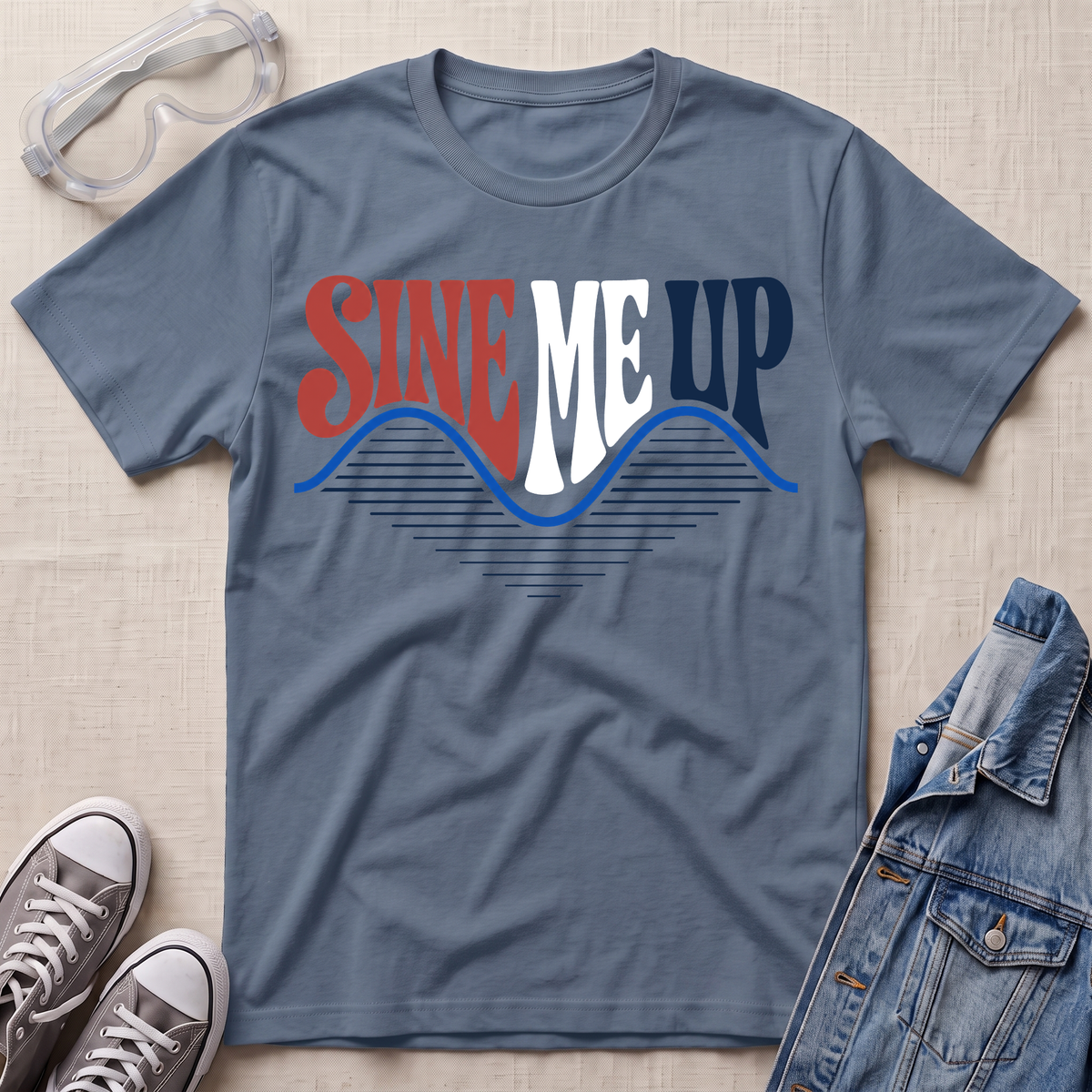 A slate blue graphic t-shirt laid flat on a light beige textured background, styled with safety goggles, gray sneakers, and a denim jacket. The t-shirt features a retro-style graphic reading 'SINE ME UP' in red, white, and blue, with a wavy blue line underneath.