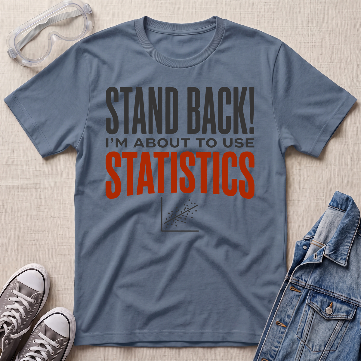 A slate blue graphic t-shirt laid flat on a light beige textured background, styled with safety goggles, gray sneakers, and a denim jacket. The t-shirt features the text 'STAND BACK! I'M ABOUT TO USE STATISTICS' in dark gray and orange-red lettering.