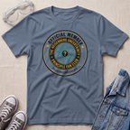 A slate blue graphic t-shirt laid flat on a light beige textured background, styled with safety goggles, gray sneakers, and a denim jacket. The t-shirt features a circular graphic that reads 'OFFICIAL MEMBER THE HEISENBERG UNCERTAINTY PRINCIPLE FAN CLUB' with a question mark atom symbol.