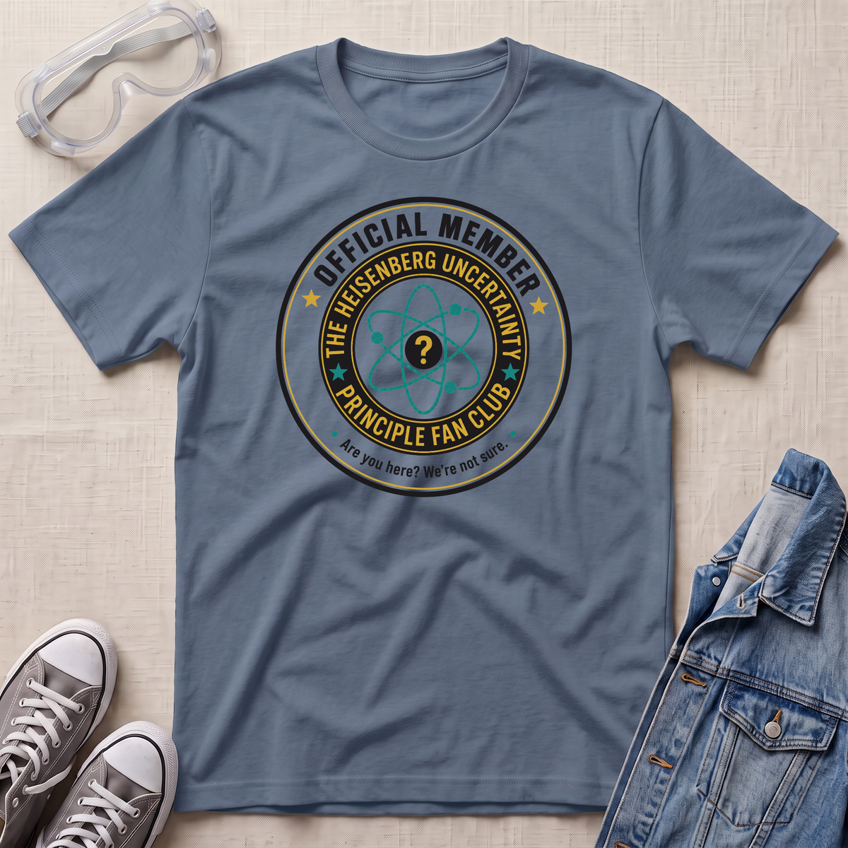 A slate blue graphic t-shirt laid flat on a light beige textured background, styled with safety goggles, gray sneakers, and a denim jacket. The t-shirt features a circular graphic that reads 'OFFICIAL MEMBER THE HEISENBERG UNCERTAINTY PRINCIPLE FAN CLUB' with a question mark atom symbol.