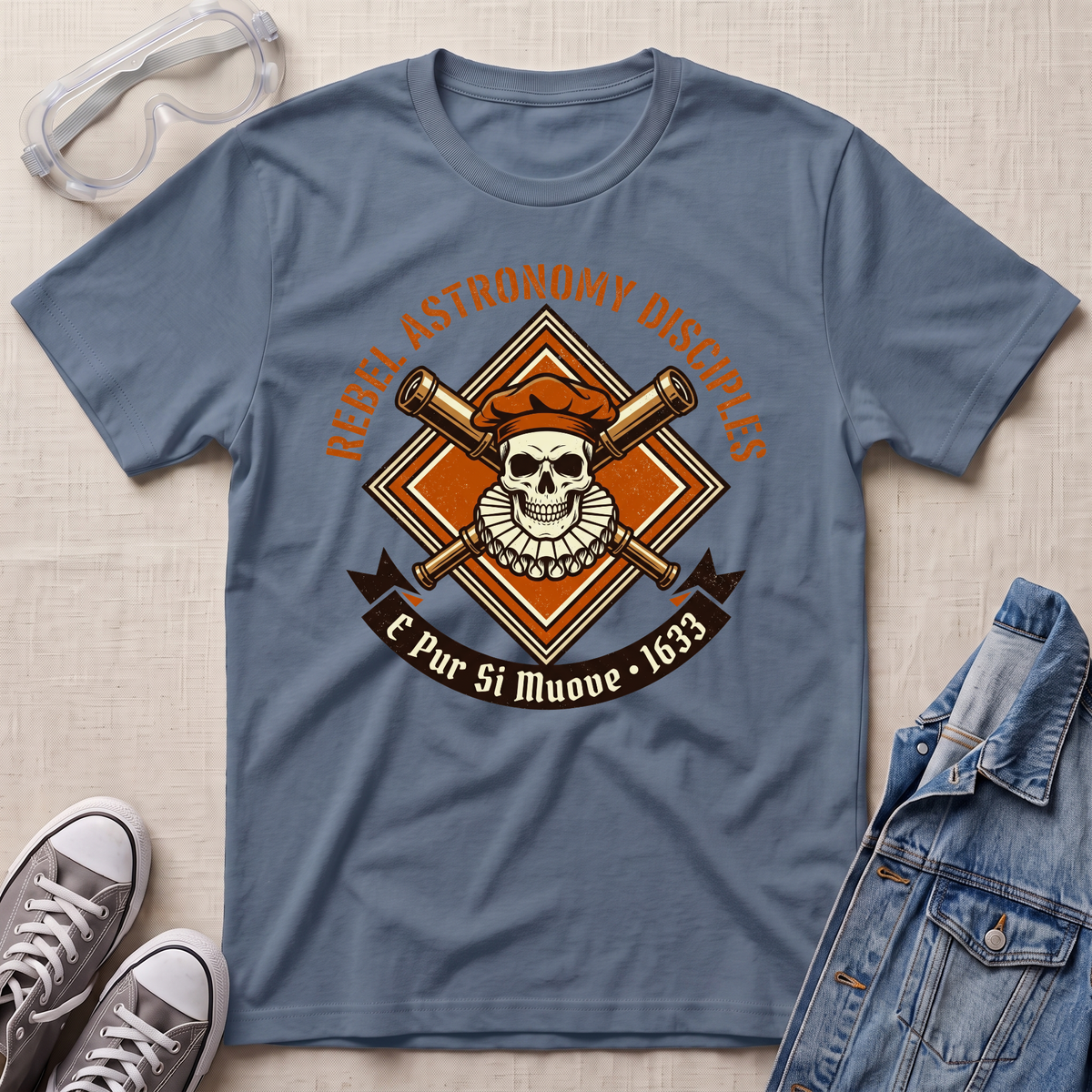 A stone blue graphic t-shirt laid flat on a light beige textured background, styled with safety goggles, gray sneakers, and a denim jacket. The t-shirt features a central graphic of a skull wearing an orange beret, crossed brass telescope barrels, and a banner reading 'REBEL ASTRONOMY DISCIPLINE' and 'E Pur Si Muove - 1633'.