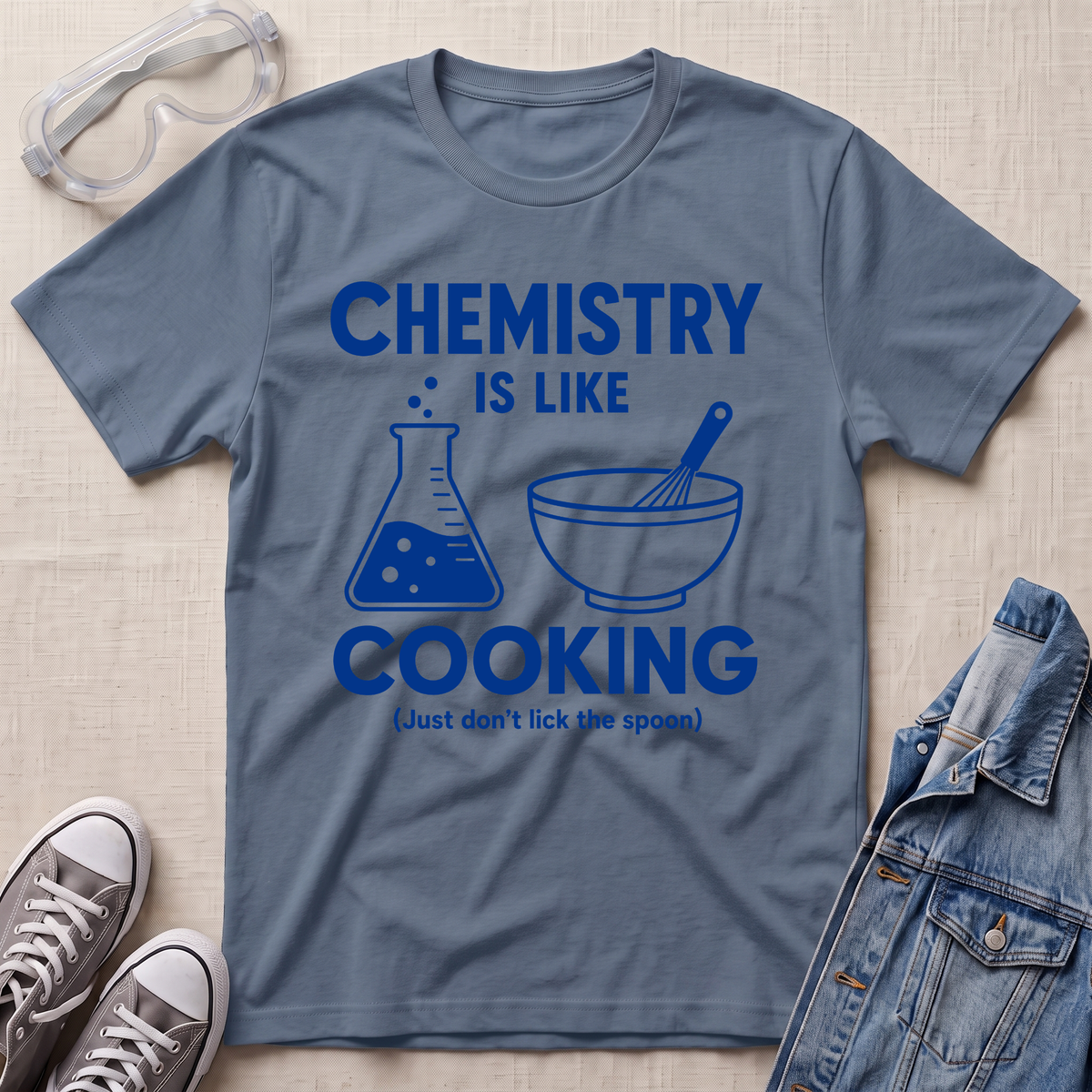 A slate blue graphic t-shirt laid flat on a beige textured background, styled with safety goggles, sneakers, and a denim jacket. The t-shirt features a blue graphic that reads 'CHEMISTRY IS LIKE COOKING (Just don't lick the spoon)' with illustrations of a beaker and a bowl with a whisk.