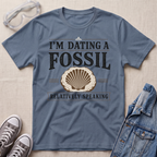 A slate blue graphic t-shirt laid flat on a textured beige background, styled with safety goggles, gray sneakers, and a denim jacket. The t-shirt features a graphic print that reads 'I'M DATING A FOSSIL RELATIVELY SPEAKING' with a seashell illustration.