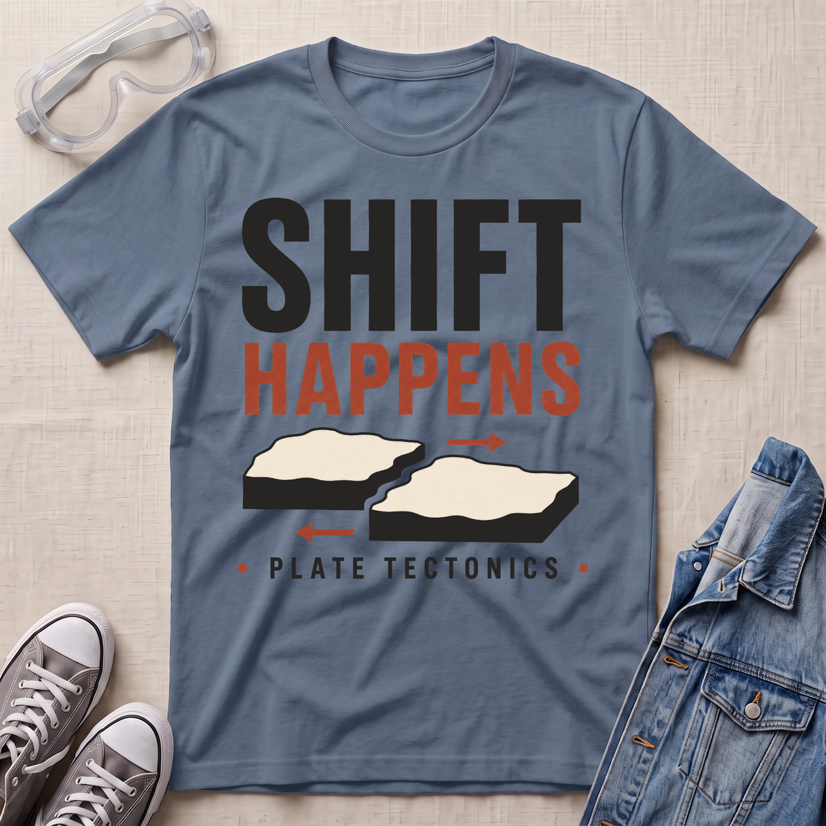 A slate blue graphic t-shirt laid flat on a light beige textured surface, styled with safety goggles, gray sneakers, and a denim jacket. The t-shirt features a graphic print that reads 'SHIFT HAPPENS' in black and orange, with two splitting landmasses and 'PLATE TECTONICS' underneath.