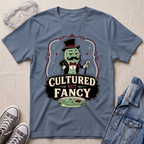 A slate blue graphic t-shirt featuring a cartoon character in a top hat and bow tie with the text 'CULTURED NOT FANCY' and a petri dish, styled in a flat lay with safety goggles, sneakers, and a denim jacket on a beige fabric background.