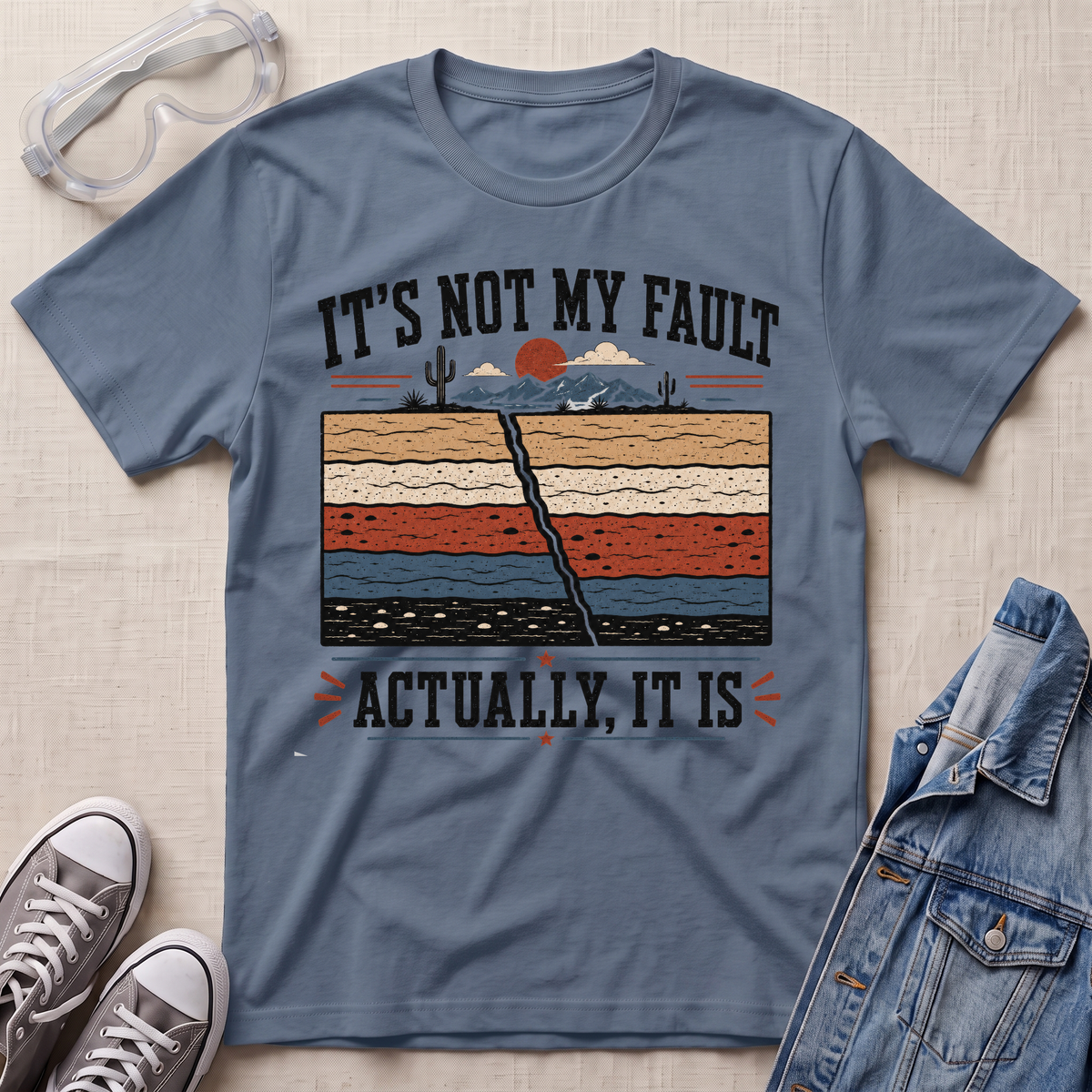 A slate blue graphic t-shirt laid flat on a light beige textured background, styled with safety glasses, gray sneakers, and a denim jacket. The t-shirt features a humorous graphic with the text 'IT'S NOT MY FAULT ACTUALLY, IT IS' surrounding a cross-section illustration of desert soil layers.
