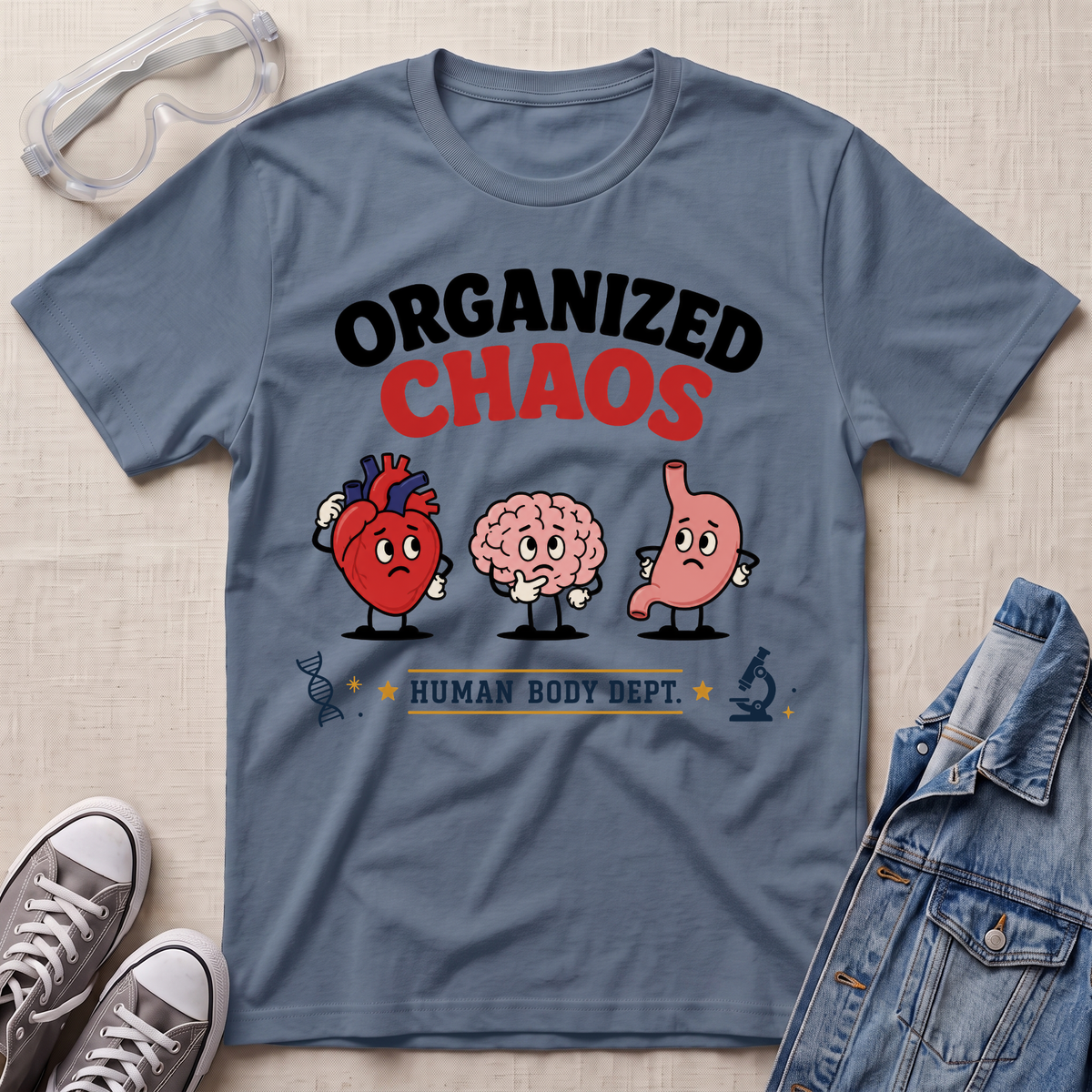 A slate blue graphic t-shirt laid flat on a light beige textured surface, surrounded by styling props including clear safety glasses, gray sneakers, and a denim jacket. The t-shirt features a cartoon graphic of a red heart, a pink brain, and a pink stomach with the text 'ORGANIZED CHAOS' and 'HUMAN BODY DEPT.