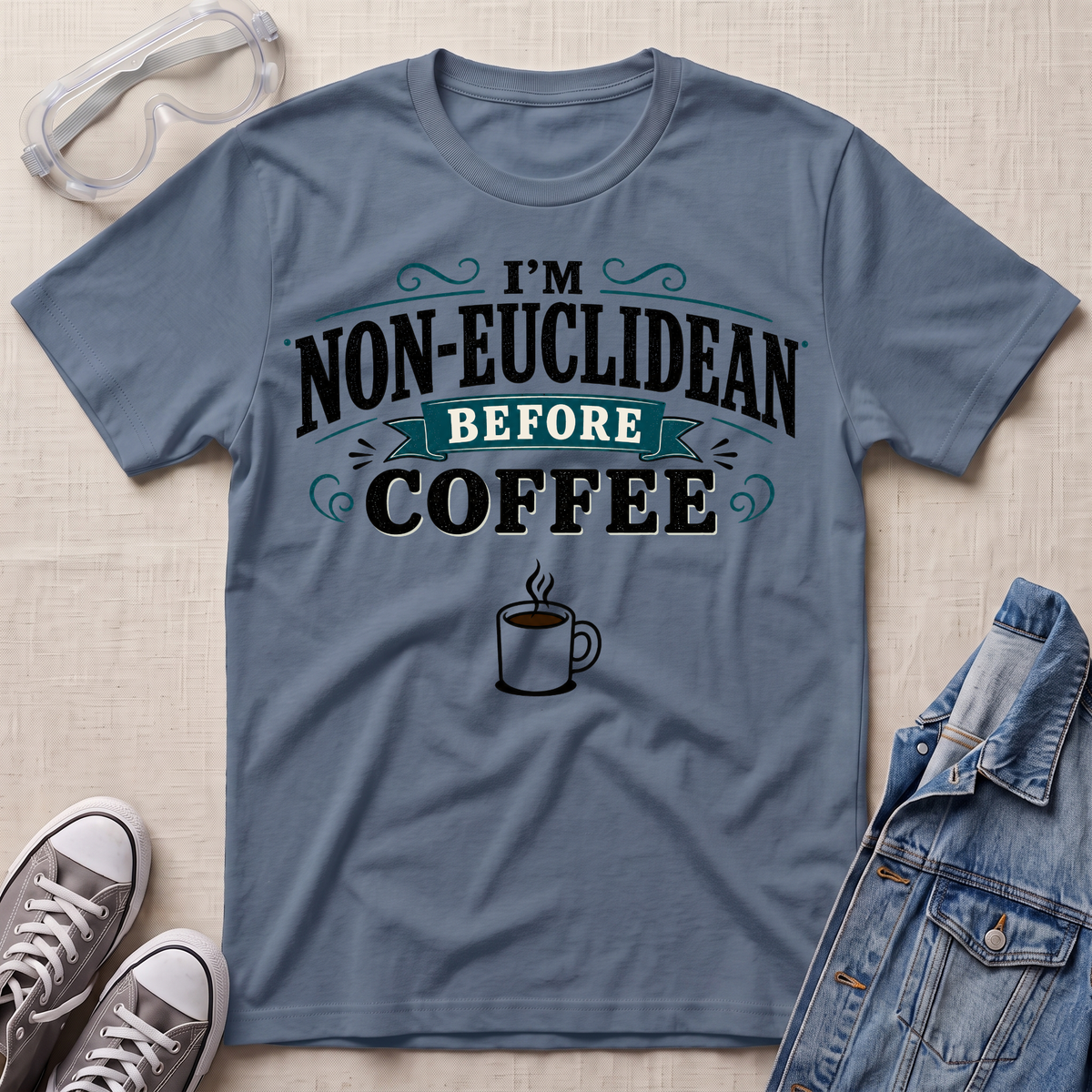 A slate blue graphic t-shirt laid flat on a beige textured background, styled with safety goggles, gray sneakers, and a denim jacket. The t-shirt features the text 'I'M NON-EUCLIDEAN BEFORE COFFEE' in black and teal lettering with a coffee cup graphic below.
