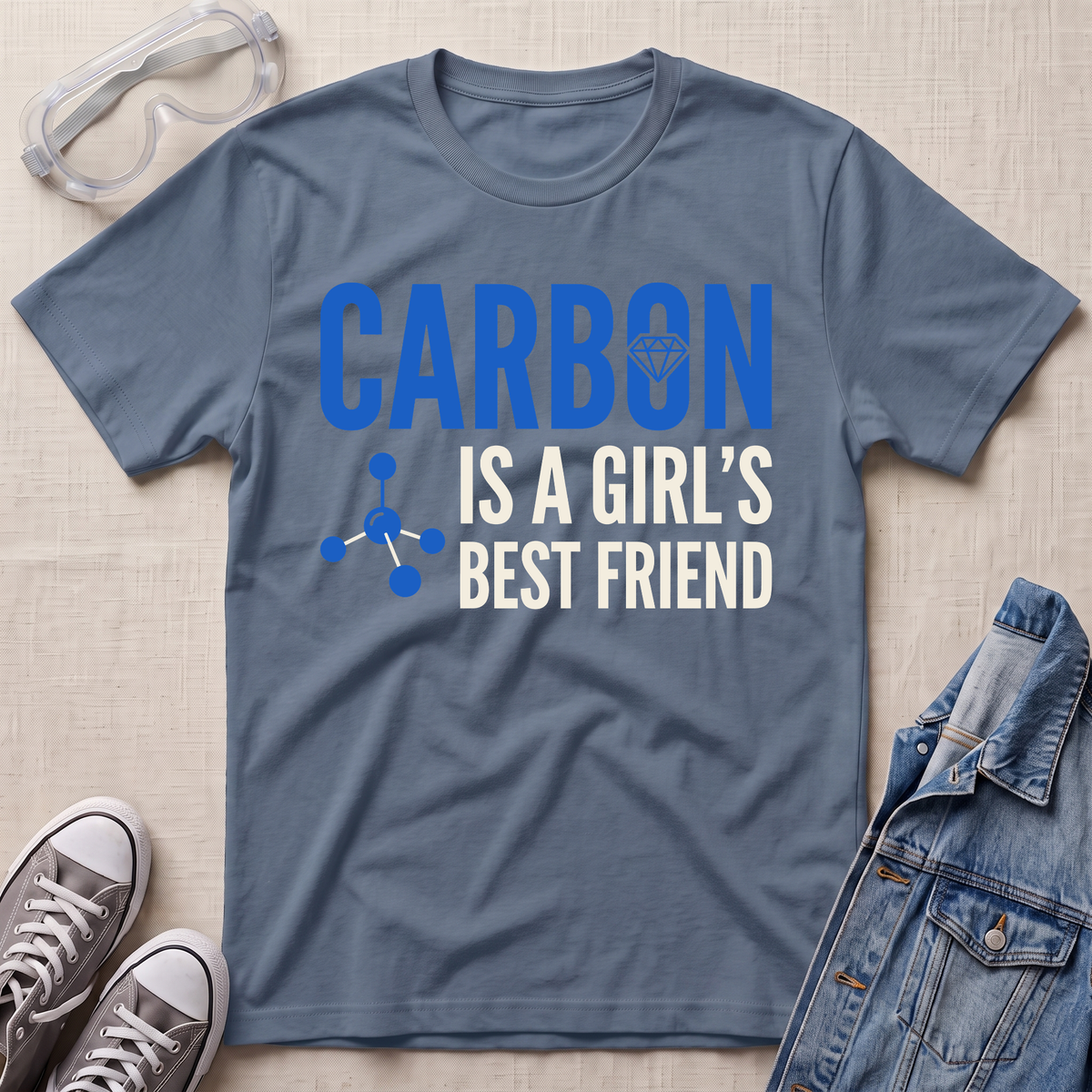 A slate blue graphic t-shirt laid flat on a light beige textured background, styled with safety goggles, gray sneakers, and a denim jacket. The t-shirt features the text 'CARBON IS A GIRL'S BEST FRIEND' in blue and white, with a small molecule graphic.