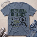 A slate blue graphic t-shirt laid flat on a light beige textured background, surrounded by styling props including safety glasses, gray sneakers, and a denim jacket. The t-shirt features a dark green and black graphic that reads 'OBSERVATIONAL BIOLOGY: I found a BUG.