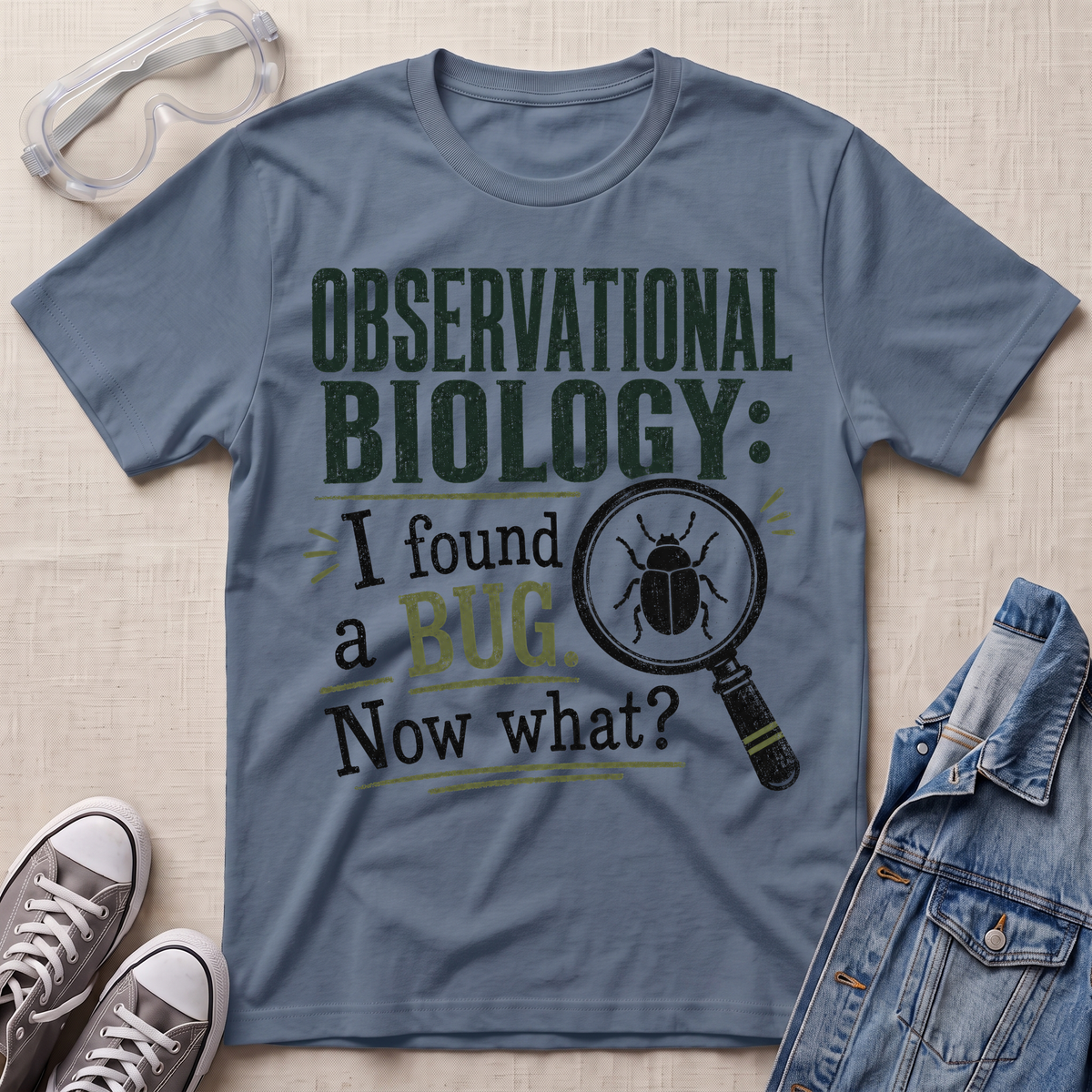 A slate blue graphic t-shirt laid flat on a light beige textured background, surrounded by styling props including safety glasses, gray sneakers, and a denim jacket. The t-shirt features a dark green and black graphic that reads 'OBSERVATIONAL BIOLOGY: I found a BUG.