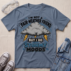 A slate blue graphic t-shirt laid flat on a light beige textured background, styled with safety goggles, gray sneakers, and a denim jacket. The t-shirt features a graphic of a storm cloud with lightning bolts and the text 'I'M NOT A FAIR-WEATHER FRIEND BUT I DO HAVE STORMY MOODS'.