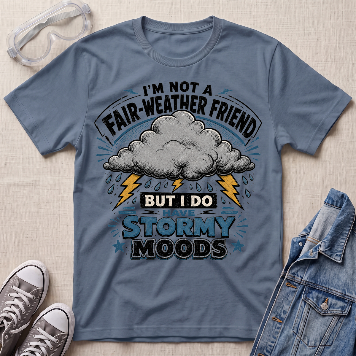 A slate blue graphic t-shirt laid flat on a light beige textured background, styled with safety goggles, gray sneakers, and a denim jacket. The t-shirt features a graphic of a storm cloud with lightning bolts and the text 'I'M NOT A FAIR-WEATHER FRIEND BUT I DO HAVE STORMY MOODS'.