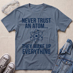A slate blue graphic t-shirt with white text reading 'NEVER TRUST AN ATOM.' and 'THEY MAKE UP EVERYTHING' with an image of an atom in between the texts.