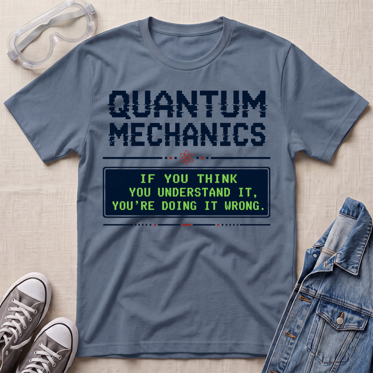 A slate blue graphic t-shirt featuring a 'Quantum Mechanics' design with a retro computer terminal screen and the text 'IF YOU THINK YOU UNDERSTAND IT, YOU'RE DOING IT WRONG.' The t-shirt is styled in a flat-lay arrangement on a beige textured background, surrounded by props including safety goggles, gray sneakers, and a denim jacket.