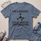 A slate blue graphic t-shirt laid flat on a light beige textured background, styled with safety goggles, gray sneakers, and a denim jacket. The t-shirt features a black printed graphic of a molecule and the text 'I AM A MOLECULE ASK ME ABOUT MY CONFORMATION'.