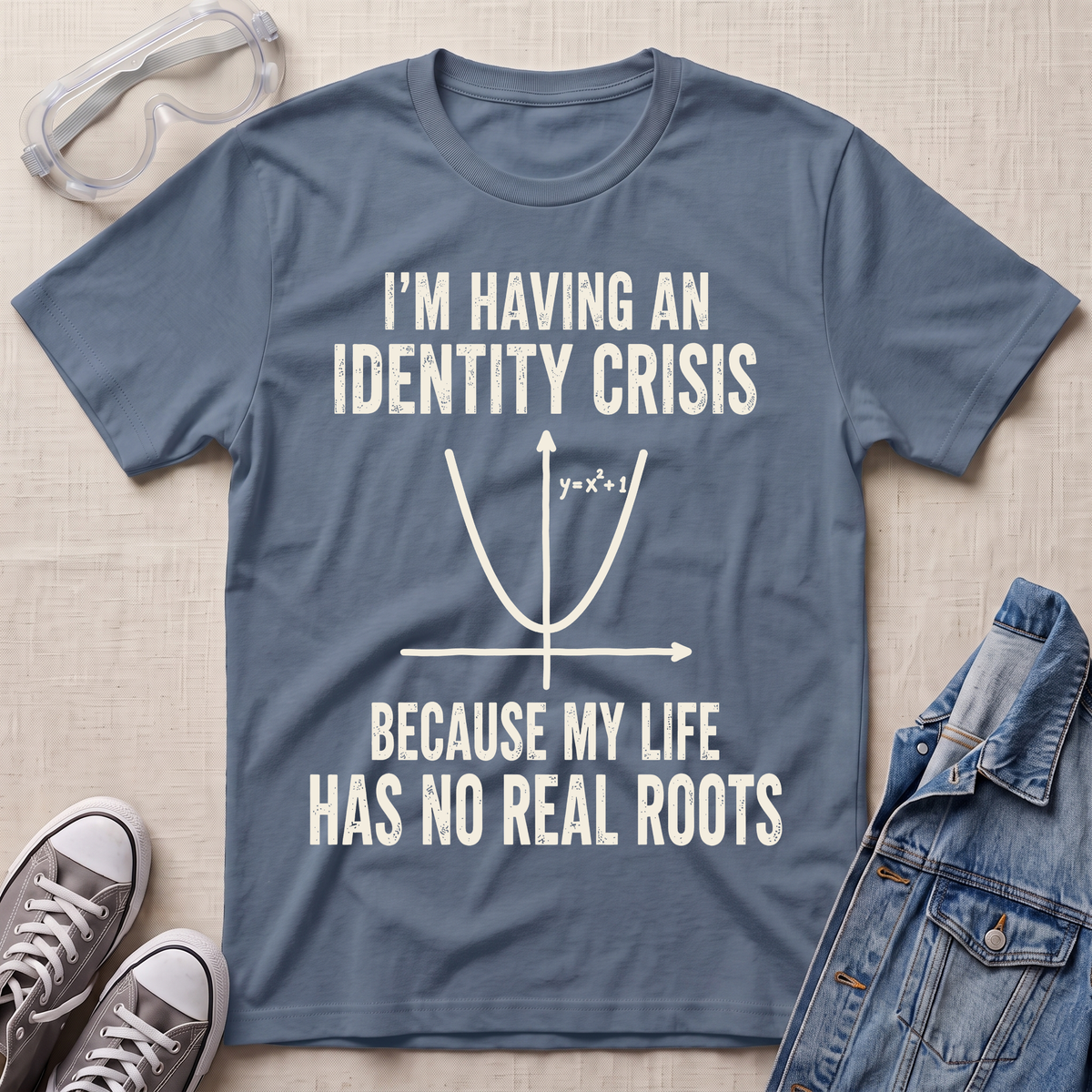 A slate blue graphic t-shirt laid flat on a light beige textured background, surrounded by styling props including safety glasses, gray sneakers, and a denim jacket. The t-shirt features a white printed graphic of a parabola with the text 'I'M HAVING AN IDENTITY CRISIS BECAUSE MY LIFE HAS NO REAL ROOTS'.