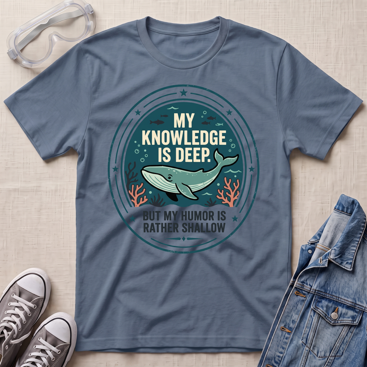 A slate blue graphic t-shirt laid flat on a light beige textured background, styled with safety goggles, gray sneakers, and a denim jacket. The t-shirt features a circular graphic with a whale and the text  'MY KNOWLEDGE IS DEEP BUT MY HUMOR IS RATHER SHALLOW'.