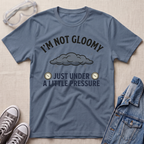 A slate blue graphic t-shirt laid flat on a light beige textured background, styled with safety goggles, gray sneakers, and a denim jacket. The t-shirt features a dark gray cloud graphic and the text 'I'M NOT GLOOMY JUST UNDER A LITTLE PRESSURE' with small clock illustrations.