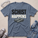 A slate blue graphic t-shirt laid flat on a beige textured background, styled with safety goggles, gray sneakers, and a denim jacket. The t-shirt features the text 'SCHIST HAPPENS' in bold black and white lettering above an illustration of a layered schist rock.