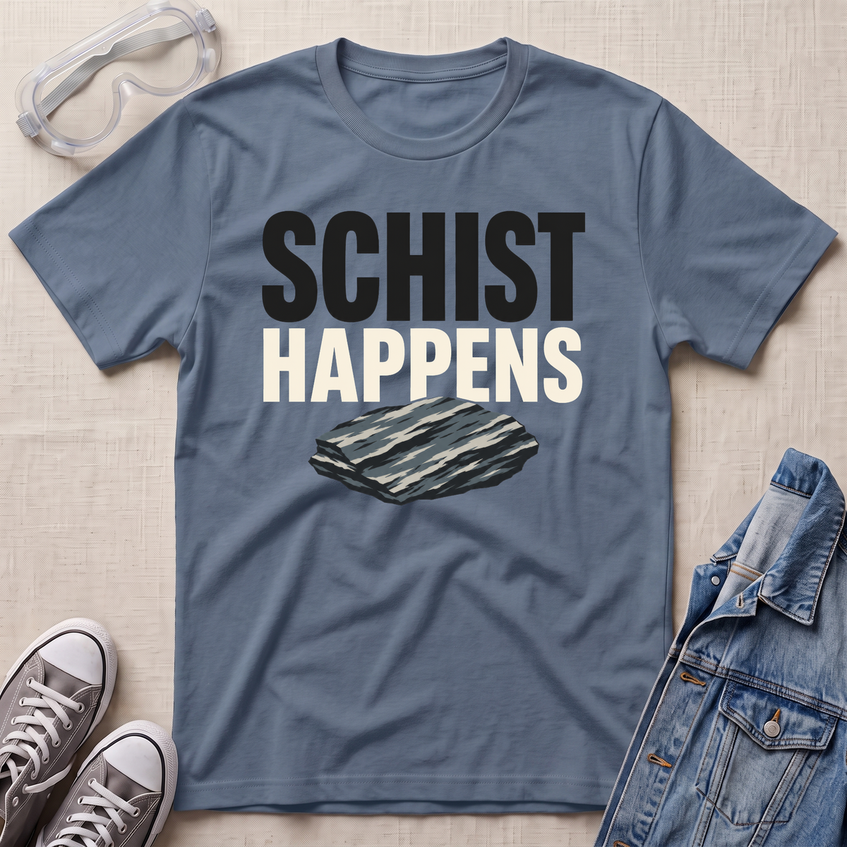 A slate blue graphic t-shirt laid flat on a beige textured background, styled with safety goggles, gray sneakers, and a denim jacket. The t-shirt features the text 'SCHIST HAPPENS' in bold black and white lettering above an illustration of a layered schist rock.