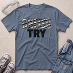A slate blue crewneck t-shirt laid flat on a beige textured background, featuring the text 'GNEISS TRY' in a distressed black and white camouflage font with 'TRY' in solid black underneath. Styled flat-lay props include a pair of clear safety goggles, gray sneakers, and a denim jacket.