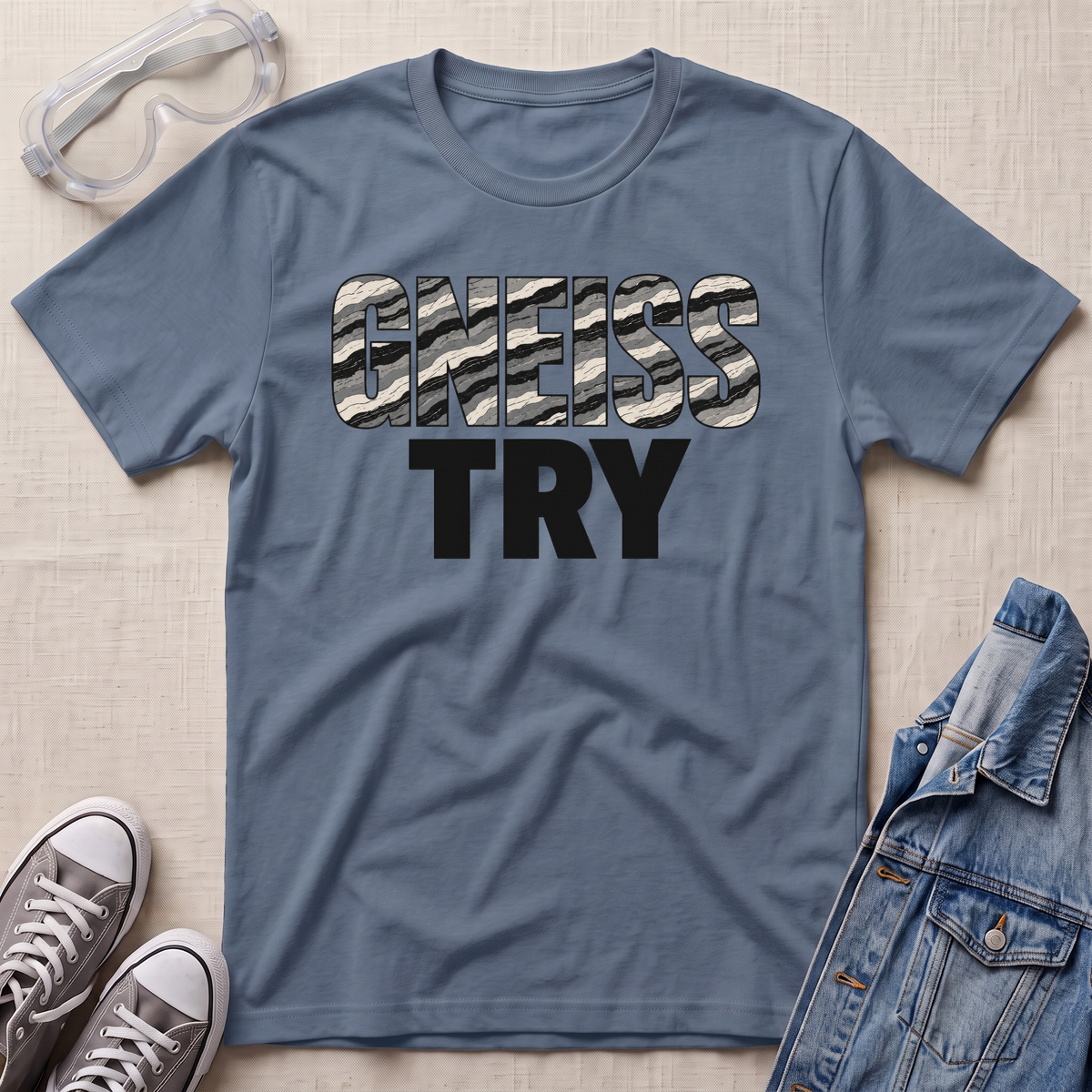 A slate blue crewneck t-shirt laid flat on a beige textured background, featuring the text 'GNEISS TRY' in a distressed black and white camouflage font with 'TRY' in solid black underneath. Styled flat-lay props include a pair of clear safety goggles, gray sneakers, and a denim jacket.