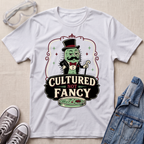 A white graphic t-shirt laid flat on a light beige fabric background, styled with clear safety goggles, gray sneakers, and a blue denim jacket. The t-shirt features a cartoon green microbe character in a top hat and bow tie above the text 'CULTURED NOT FANCY' with a petri dish graphic.