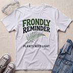 A white graphic t-shirt laid flat on a light beige textured background, styled with safety goggles, gray sneakers, and a denim jacket. The t-shirt features a green and black graphic that reads 'FRONDLY REMINDER' with a fern leaf and 'PLANTS NEED LIGHT'.