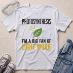 A white graphic t-shirt laid flat on a light beige textured background, surrounded by styling props including clear safety goggles, gray sneakers, and a blue denim jacket. The t-shirt features a green leaf graphic and the text 'PHOTOSYNTHESIS I'M A BIG FAN OF LIGHT WORK' in black and yellow.