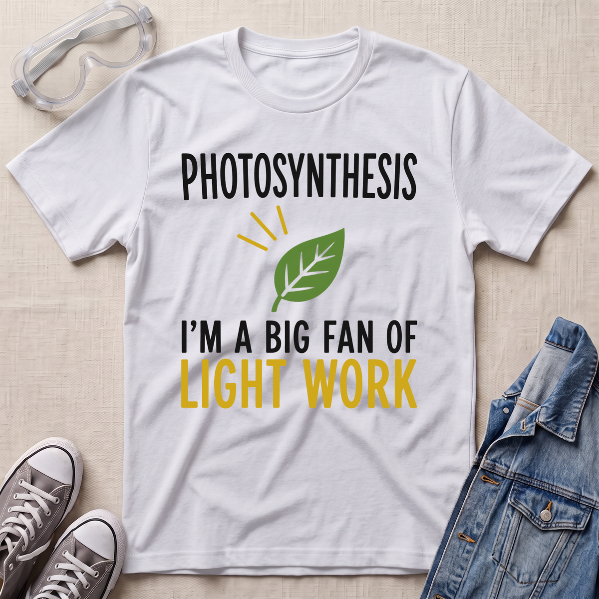 A white graphic t-shirt laid flat on a light beige textured background, surrounded by styling props including clear safety goggles, gray sneakers, and a blue denim jacket. The t-shirt features a green leaf graphic and the text 'PHOTOSYNTHESIS I'M A BIG FAN OF LIGHT WORK' in black and yellow.
