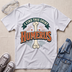 A white graphic t-shirt laid flat on a light wood surface, featuring a bone graphic and the text 'I FIND THIS SHIRT HUMERUS' in orange and green. The shirt is styled with safety glasses, gray sneakers, and a denim jacket.