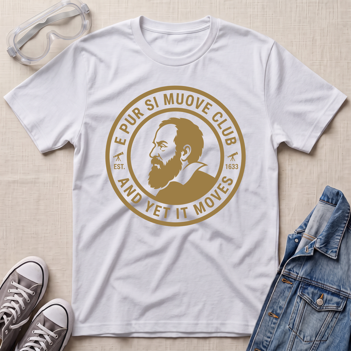 A white short-sleeve graphic T-shirt laid flat on a light beige textured surface, surrounded by styling props including clear safety goggles, gray sneakers, and a blue denim jacket. The shirt features a large circular tan/gold print with a portrait and the text 'E PUR SI MUOVE CLUB AND YET IT MOVES'.