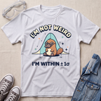 A white graphic t-shirt featuring a platypus wearing glasses under a blue bell curve with the text 'I'M NOT WEIRD' and 'I'M WITHIN ±1σ'. The t-shirt is styled in a flat-lay arrangement on a textured beige background, surrounded by props including safety goggles, gray sneakers, and a denim jacket.