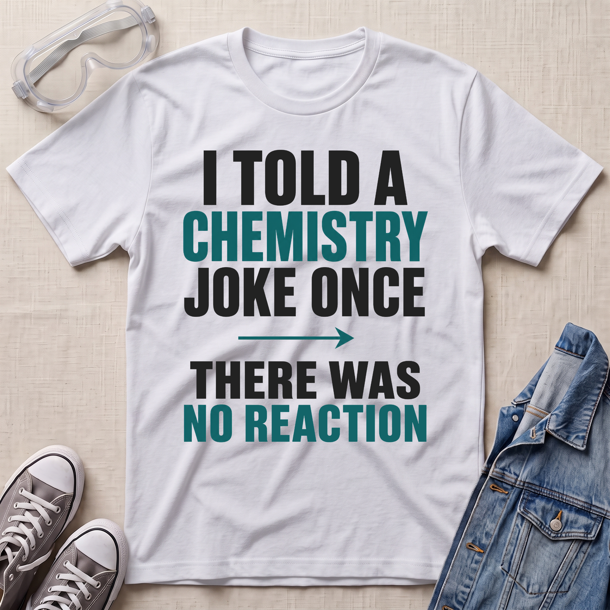 A white graphic t-shirt laid flat on a light beige textured background, surrounded by styling props including safety goggles, gray sneakers, and a denim jacket. The t-shirt features a printed slogan in black and teal text that reads 'I TOLD A CHEMISTRY JOKE ONCE THERE WAS NO REACTION'.