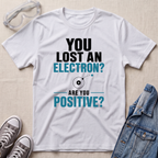A white graphic t-shirt laid flat on a light beige textured surface, surrounded by styling props including safety goggles, gray sneakers, and a blue denim jacket. The t-shirt features a printed design that reads 'YOU LOST AN ELECTRON? ARE YOU POSITIVE?' in black and teal text with a simple atom illustration.