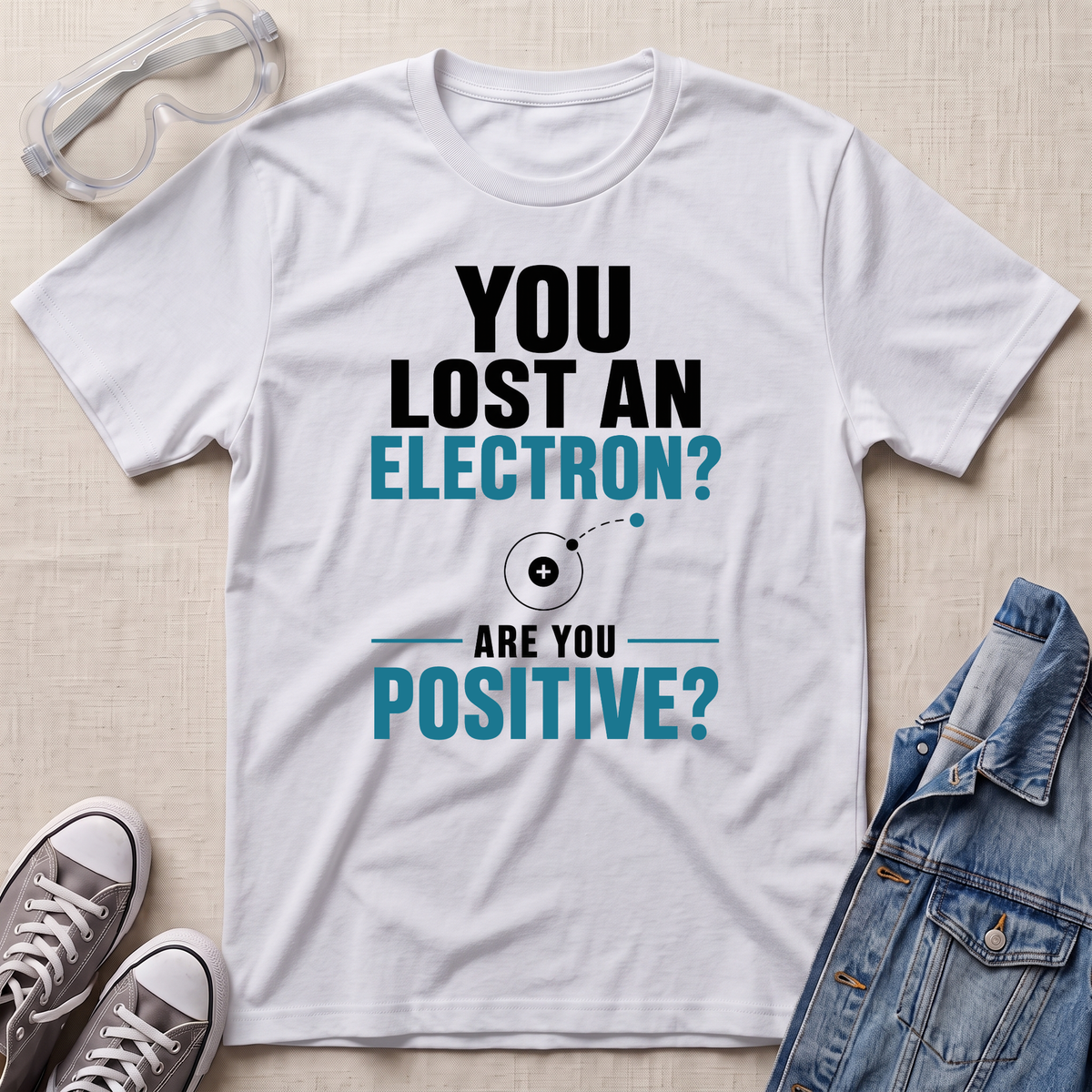 A white graphic t-shirt laid flat on a light beige textured surface, surrounded by styling props including safety goggles, gray sneakers, and a blue denim jacket. The t-shirt features a printed design that reads 'YOU LOST AN ELECTRON? ARE YOU POSITIVE?' in black and teal text with a simple atom illustration.