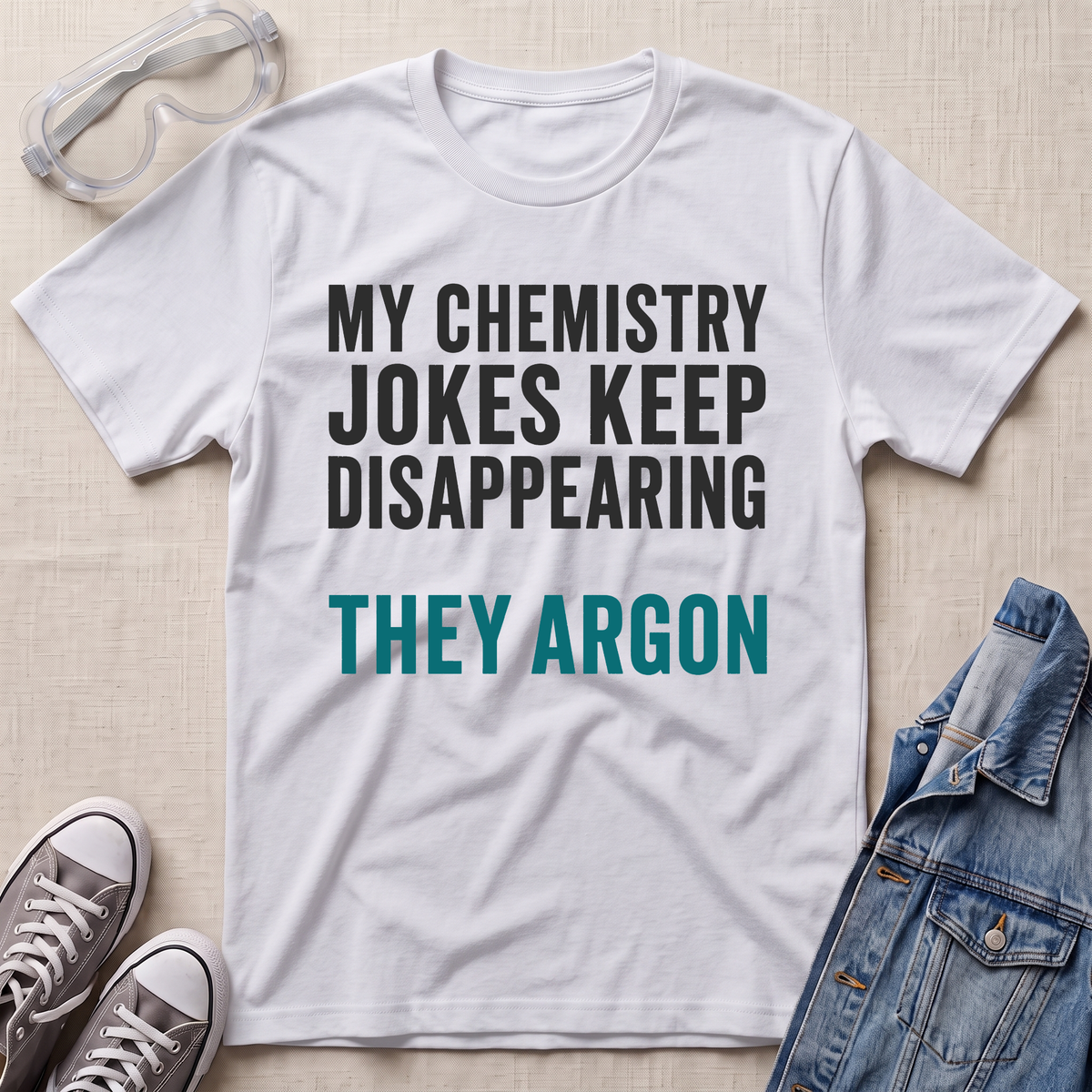 A white graphic t-shirt laid flat on a light beige textured background, surrounded by styling props including safety goggles, gray sneakers, and a denim jacket. The t-shirt features the text 'MY CHEMISTRY JOKES KEEP DISAPPEARING THEY ARGON' in black and teal lettering.