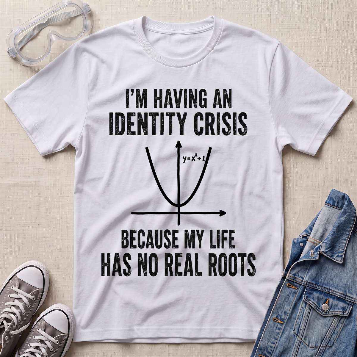 A white graphic t-shirt laid flat on a light beige fabric background, styled with clear safety glasses, gray sneakers, and a blue denim jacket. The t-shirt features a black printed graphic of a parabola with the text 'I'M HAVING AN IDENTITY CRISIS BECAUSE MY LIFE HAS NO REAL ROOTS'.