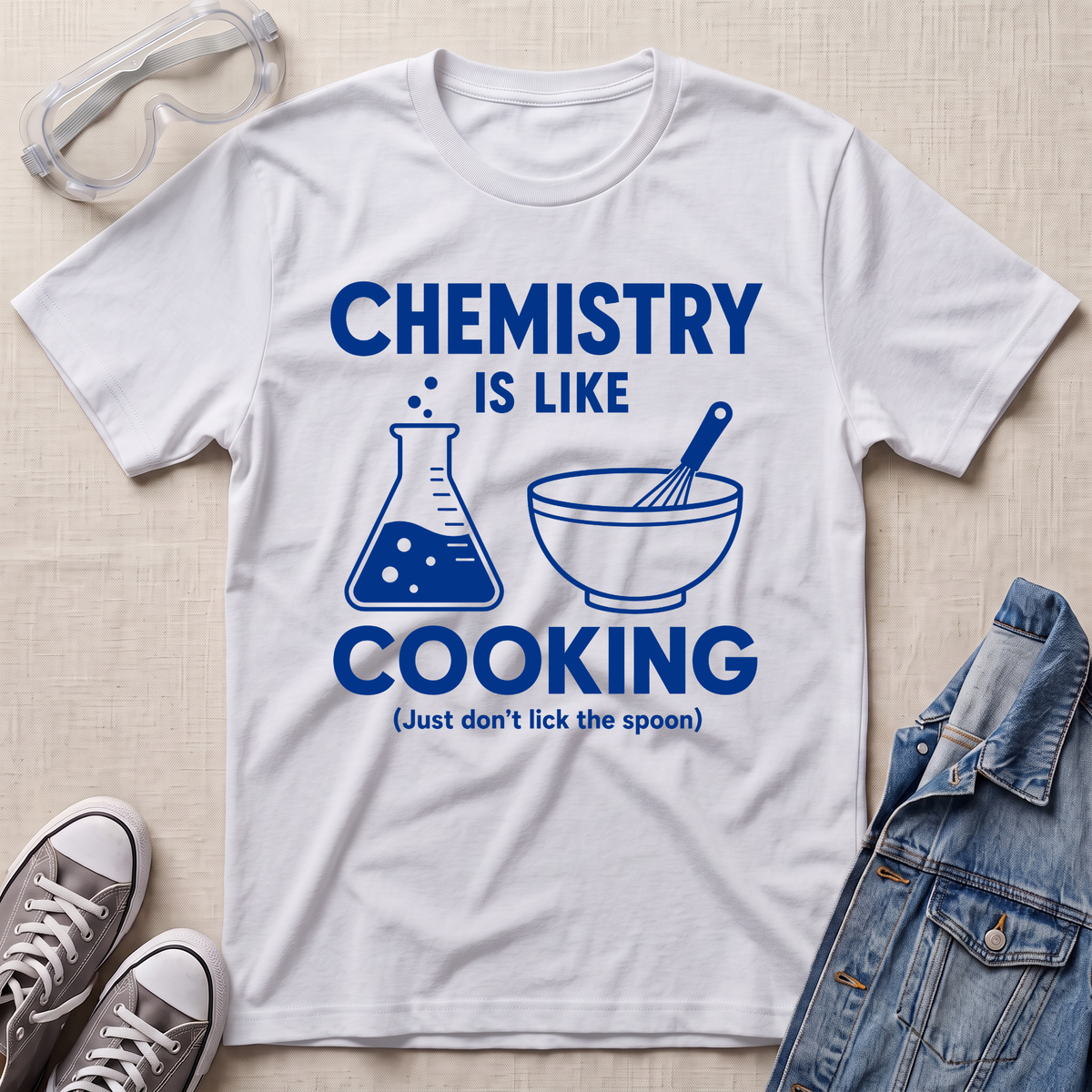 A white short-sleeve graphic T-shirt laid flat on a light beige fabric background, styled with safety goggles, gray sneakers, and a denim jacket. The shirt has a blue printed design that reads “CHEMISTRY IS LIKE COOKING (Just don’t lick the spoon)” with a beaker and bowl-with-whisker illustration.
