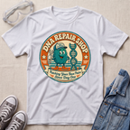 A white short-sleeve graphic T-shirt laid flat on a light fabric surface, surrounded by styling props including clear safety glasses, gray sneakers, and a blue denim jacket. The shirt features a large circular retro-style graphic reading 'DNA REPAIR SHOP' with a cartoon teal repair character and a DNA double helix.