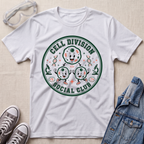 A white short-sleeve graphic T-shirt laid flat on a light fabric surface, surrounded by styling props including clear safety goggles, gray sneakers, and a blue denim jacket. The shirt has a large circular green and orange cartoon graphic reading 'CELL DIVISION SOCIAL CLUB' with illustrated cells and small scientific icons.