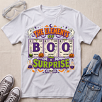 A white graphic T-shirt laid flat on a light fabric background, styled with clear safety goggles, gray sneakers, and a denim jacket. The shirt features a colorful Halloween-themed design reading 'THE ELEMENTS OF BOO SURPRISE' with periodic-table-style elements, ghosts, pumpkins, bats, and purple decorative accents.