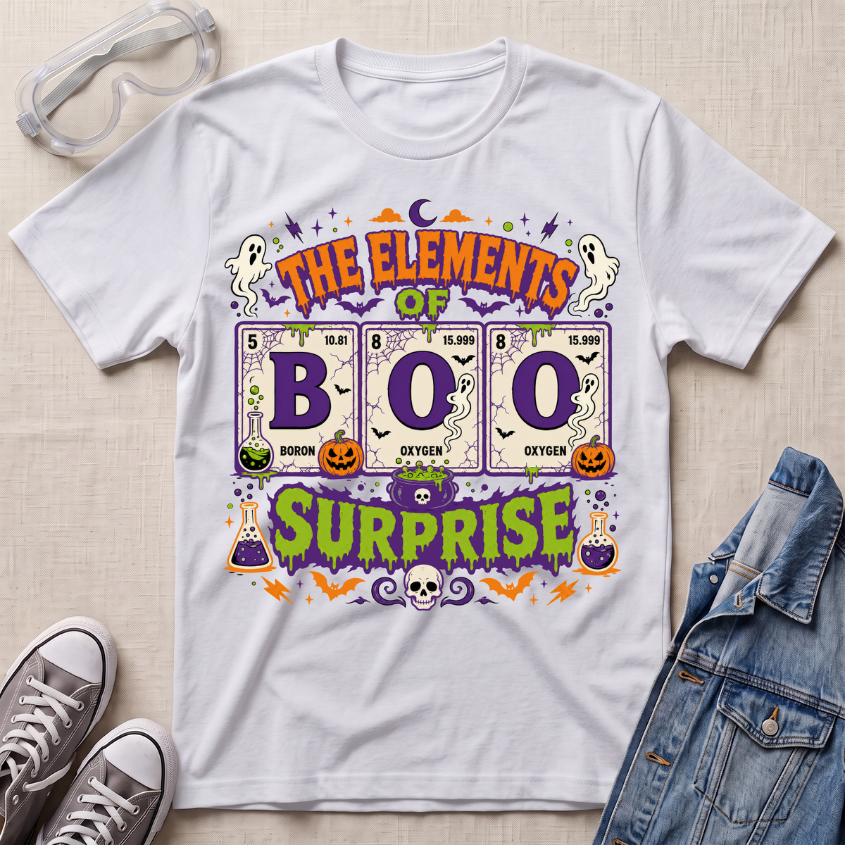 A white graphic T-shirt laid flat on a light fabric background, styled with clear safety goggles, gray sneakers, and a denim jacket. The shirt features a colorful Halloween-themed design reading 'THE ELEMENTS OF BOO SURPRISE' with periodic-table-style elements, ghosts, pumpkins, bats, and purple decorative accents.