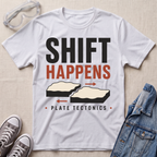 A white graphic t-shirt laid flat on a light beige textured background, styled with safety goggles, gray sneakers, and a denim jacket. The t-shirt features a retro-style graphic that reads 'SHIFT HAPPENS' in black and orange-red text, with illustrations of tectonic plates and the words 'PLATE TECTONICS'.